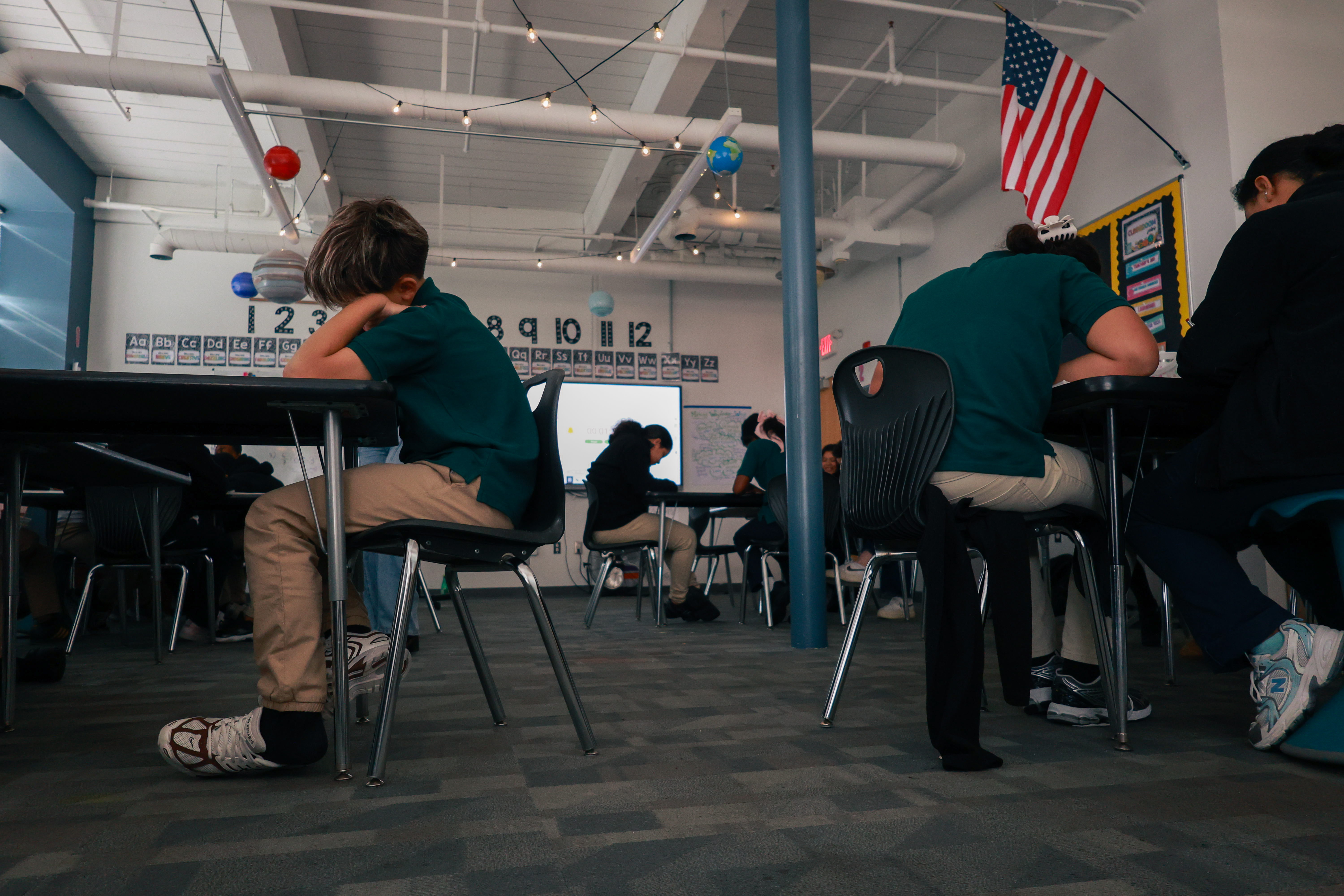 Photo of students in math class. One student leans his head into his hand as he looks at his paper. Other students throughout the room similarly lean over their work intently.